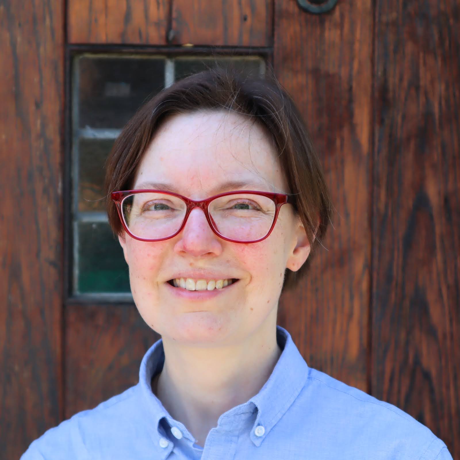 Jennifer Holt with red glasses standing in front of a brown wooden door