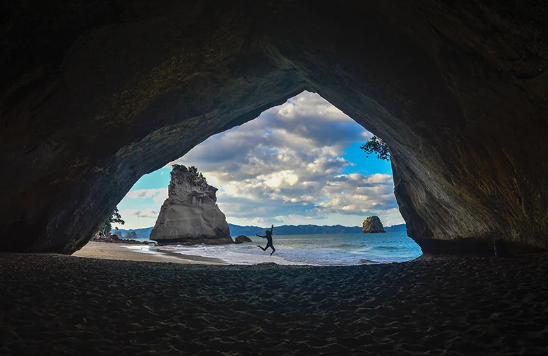 A student jumping on a beach, seen from a cave
