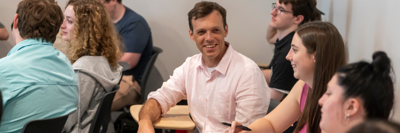 People interacting in a classroom setting, with a man in a pink shirt smiling.