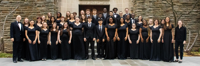 A choir group in formal black attire, standing in rows in front of a stone building.