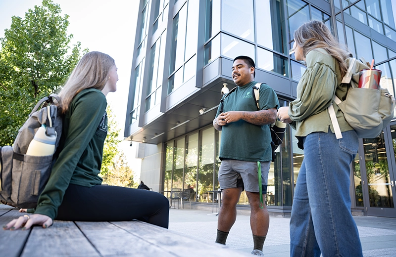 Three students gather to chat outside by the large glass facade of the Fernandez Center