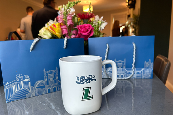 A coffee mug featuring the Newcastle and Loyola logos sits on a table with Newcastle University gift bags and a floral arrangement in the background