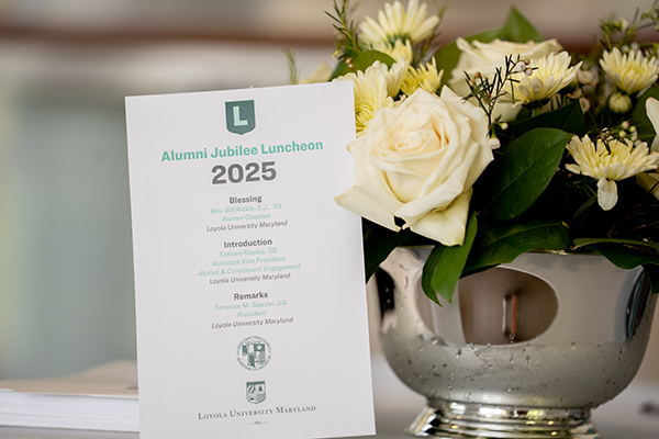 An event card for the 2025 Alumni Jubilee Luncheon posed next to a white rose centerpiece