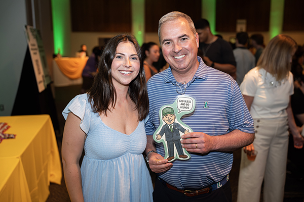 A senior poses for a photo with Loyola President Terrence M. Sawyer, J.D., who is holding a cartoon image of himself with a speech bubble that reads "God bless and Go Hounds!"