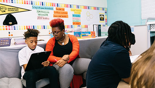 Teacher on a couch in a classroom with a student while they do work together on a laptop