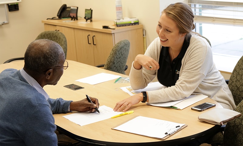 A man writing on paper while sitting in front of a Clinical Centers staff member