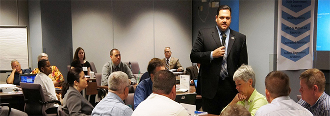 A trainer holding a microphone addresses an audience at circular tables