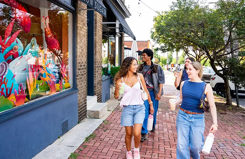 Four students chat walk down a brick sidewalk in Baltimore while holding drinks and passing a colorful storefront