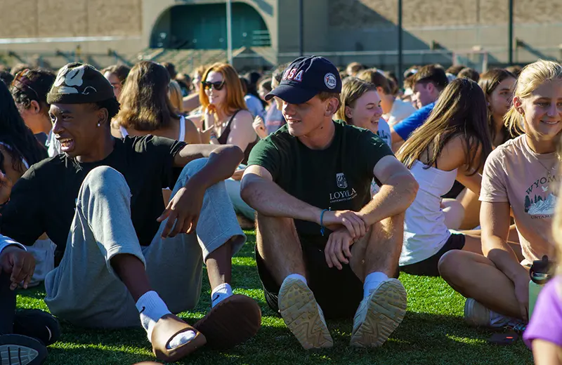 The students of the first-year class sit, laugh, and socialize while sitting on the turf field on Loyola's campus