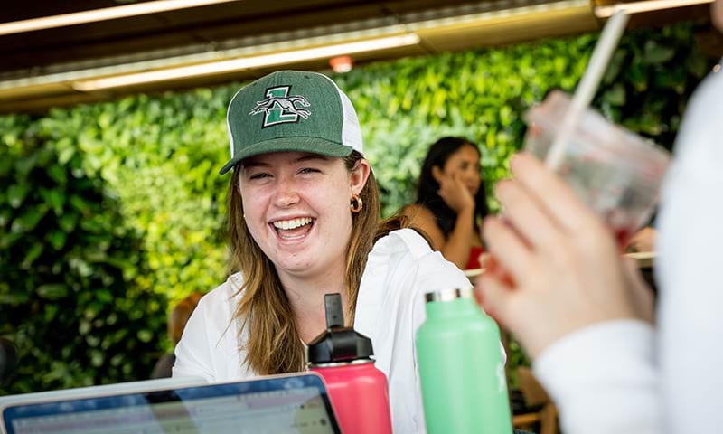 A female student laughing while wearing a Loyola baseball cap and chatting with classmates