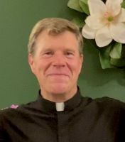 Headshot of Father Stephen Spahn, S.J. smiling with a green background and white flower in upper right corner