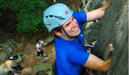 Student in a helmet climbing up an outdoor wall of rock while a student spots him from the ground