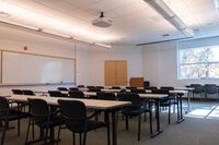 Classroom with rows of tables, podium, whiteboard, and window looking out to the wooded surrounding area