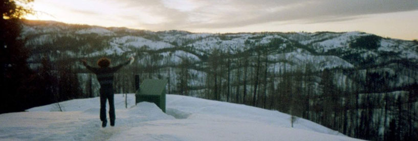 Student in snow clothes at the top of a snowy mountain jumping with arms raised and back to camera