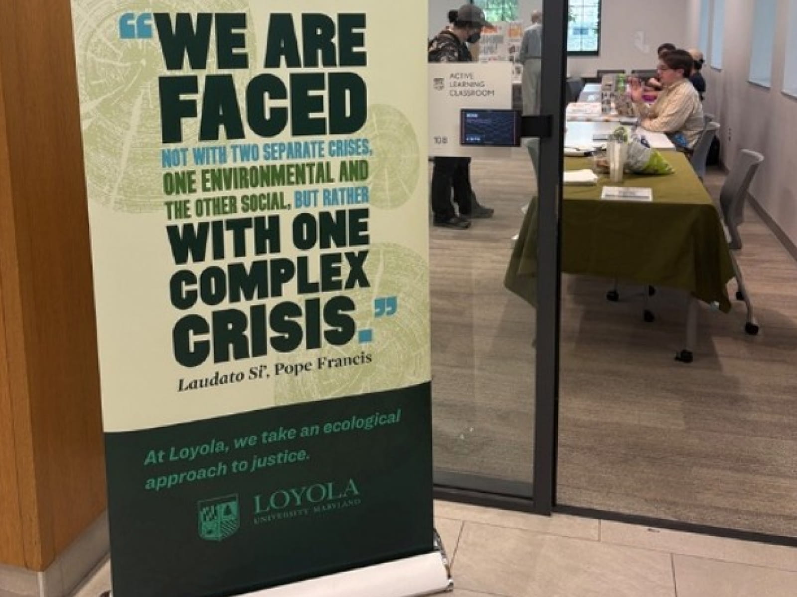 standing banner outside of a classroom that reads We are faced with one complex crisis and the loyola logo