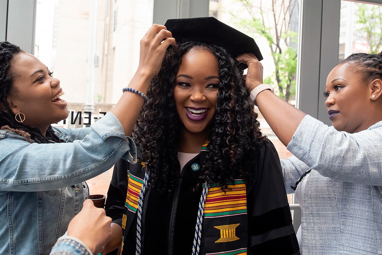 A graduating student smiles as two people pin a commencement hat on her head