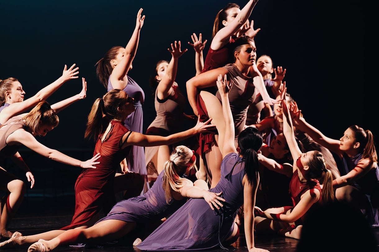 Loyola dancers posing together in a large group, wearing red, purple, and brown dresses in front of a black background