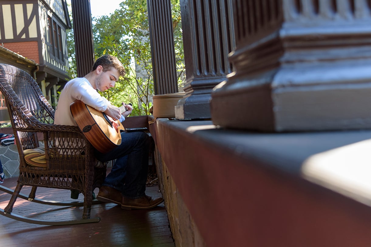 A student playing a guitar while sitting on a rocking chair on the Humanities building porch