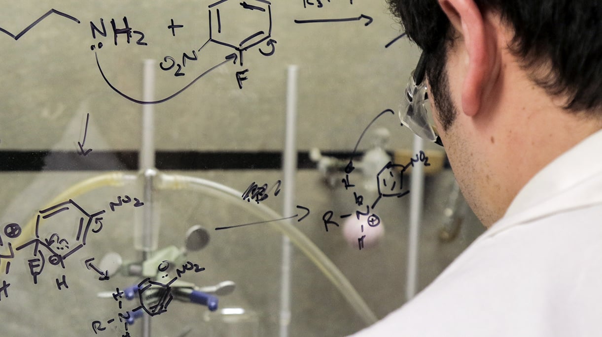 A chemist looking at a glass window with chemistry notes written on it in dry erase marker