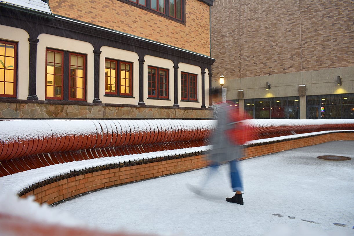 A blurred student is seen walking through the snow behind the Humanities building