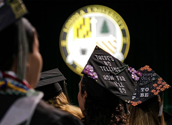 Students wearing decorated graduation caps looking at a stage with the Loyola seal in the background