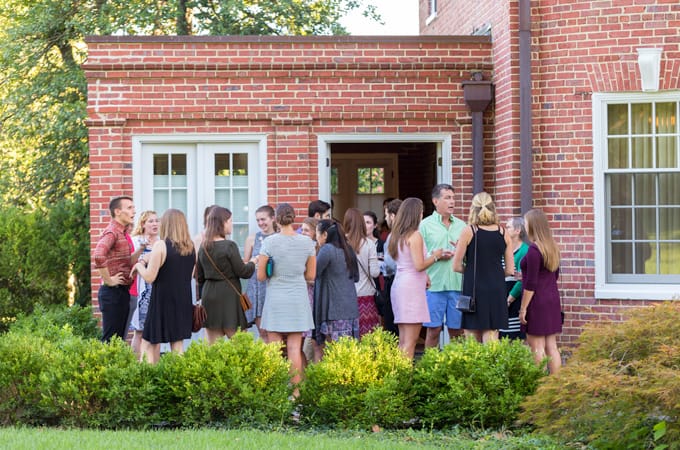 President Linnane and a group of students casually talking on the back porch of the red-bricked alumni house