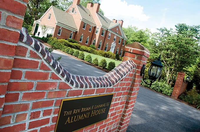 A red brick wall with a sign reading The Rev. Brian F. Linnane Alumni House, and a red-bricked building in the background