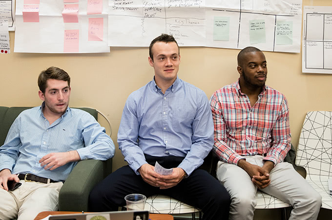 Three students sitting in chairs in front of brainstorm papers taped to the wall behind them