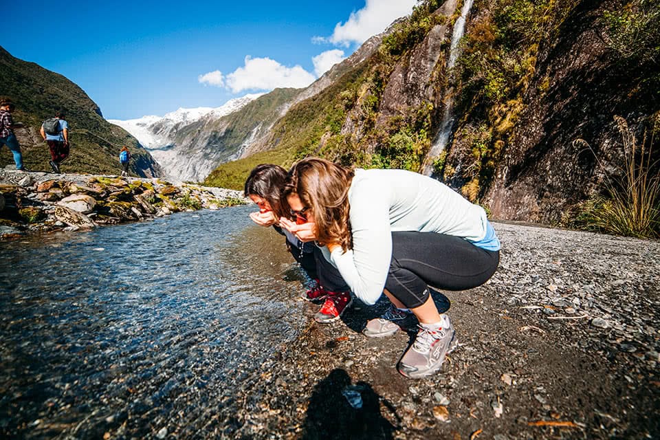 Two students crouching and drinking from a stream