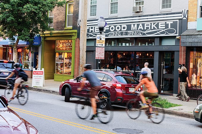 People riding bikes down a street, passing shops and parked cars