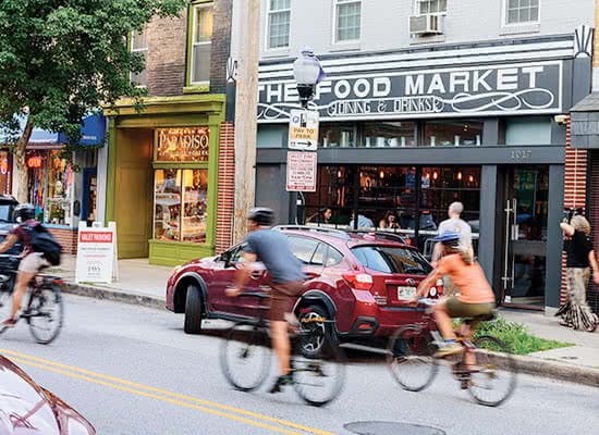 People riding bikes down a street, passing shops and parked cars