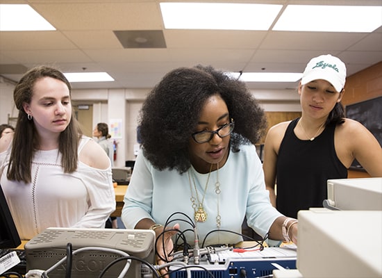 Two students watching over the shoulder of a teacher working with engineering equipment