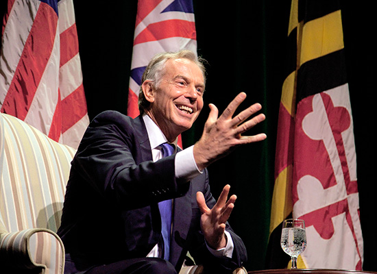 Tony Blair sits and speaks on stage with the US, UK, and Maryland flags behind him.