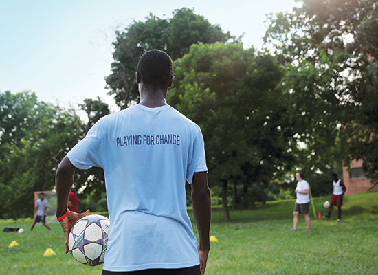 Young student stands holding a soccer ball. The back of his shirt reads 