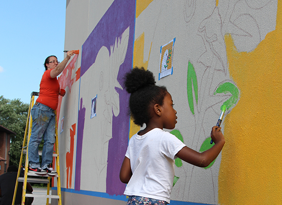 Young girl paints bright green leaves on outdoor mural.