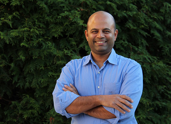 Ravi Srinivasan smiles and crosses his arms in photo with evergreen trees behind him.