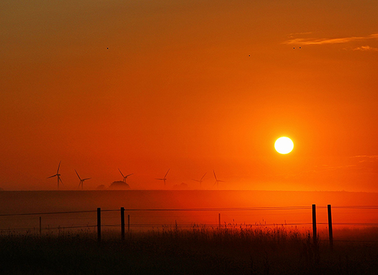 Red sunset over a field with wind turbines on the hazy horizon.