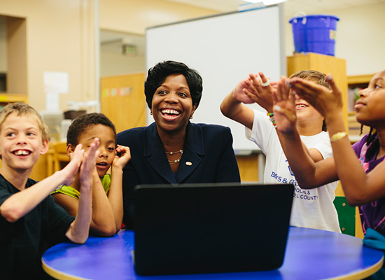 Photo of Kimberly Ellison-Taylor sitting at a desk with four of her students, smiling and laughing.