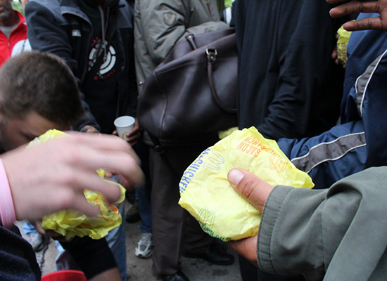 Amidst people, a hand holds a McDonalds' sandwich.