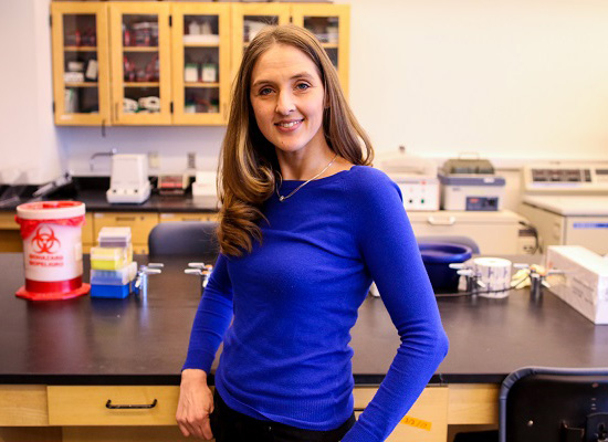 Birgit poses by large lab desk. smiling.