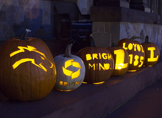 Carved pumpkins on the humanities manor porch with Loyola symbols and logos glowing with candlelight.