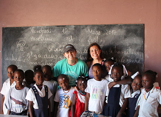 Two Loyola students stand with Haitian schoolchildren in front of a blackboard.