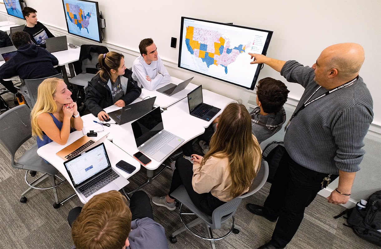 Several students sitting at a table watching a professor pointing at a screen above them