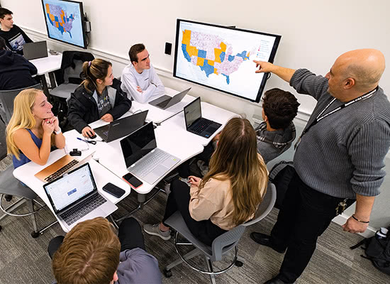 Several students sitting at a table watching a professor pointing at a screen above them