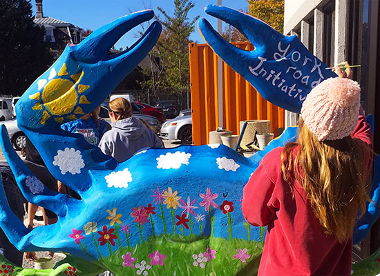 Students painting a large statue of a crab with bright colors that read 'York Road Initiative' outside on a sunny day