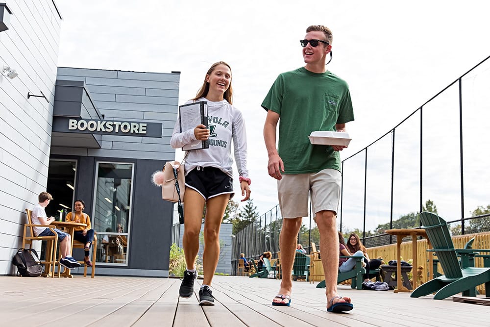 Two students walking on a wood deck, with the bookstore in the background, and other students sitting on chairs outdoors