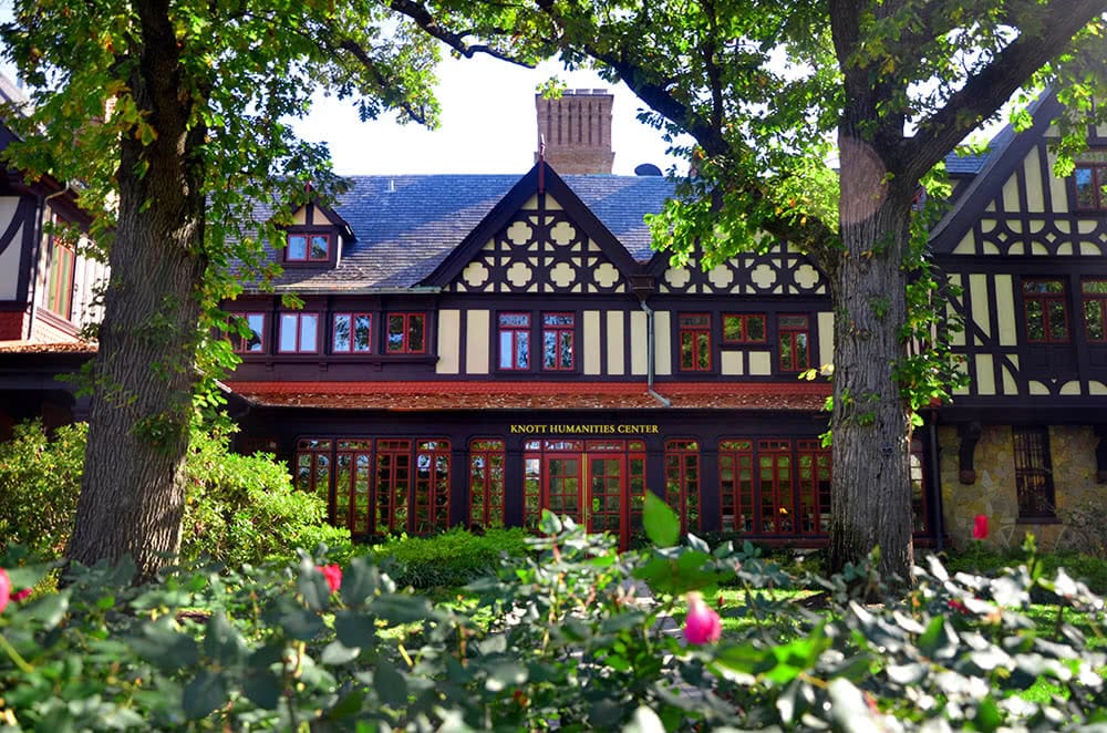 The front of the Humanities Building framed by two large trees and rosebushes with red flowers