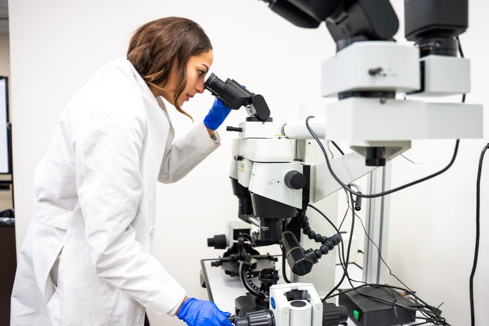 A student in a lab coat and latex gloves looking through a microscope.