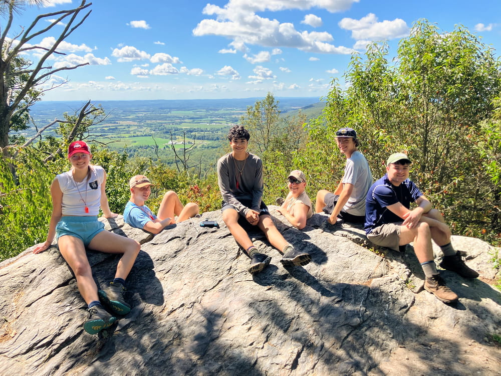 A group of students sitting on a large boulder enjoying the view from a mountaintop during a hike.