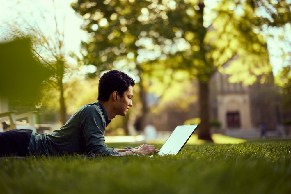 A student lying in the grass and looking at his laptop.